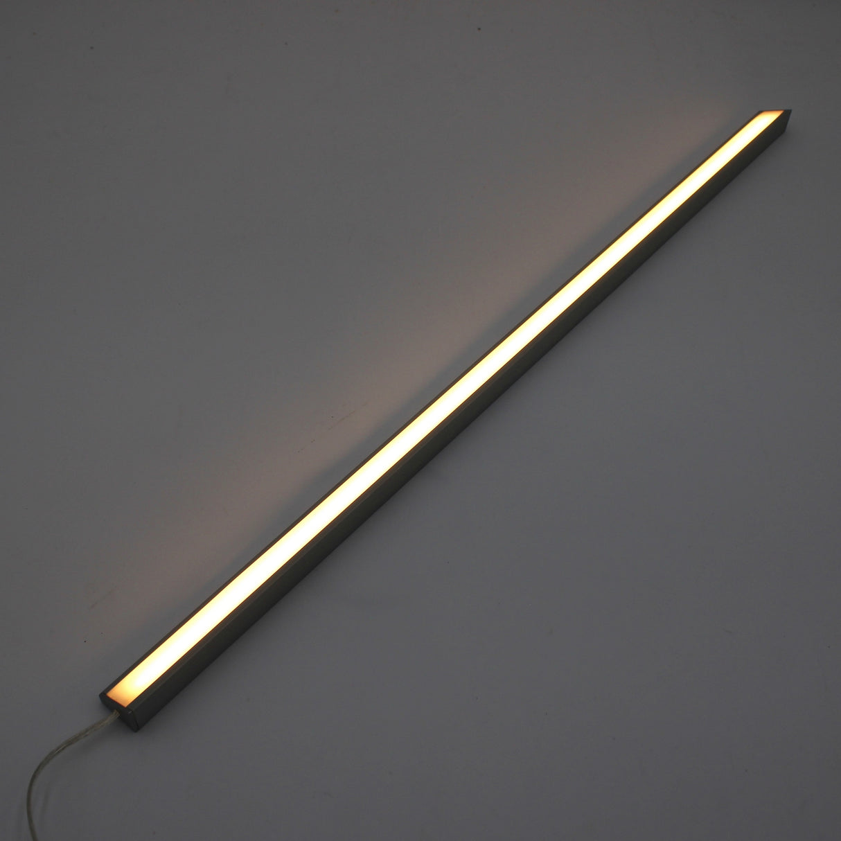 The White Universal Surface-mount LED Corner Lighting Fixture (24V) ~ Corner14 Series features a minimalist, modern design and emits a soft, warm glow when mounted diagonally against a plain gray wall.