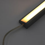 A close-up of the White Universal Surface-mount LED Corner Lighting Fixture (24V) ~ Corner14 Series, featuring a warm white glow in a dark metal frame with a transparent power cord, ideal for under cabinet lighting against a plain background.