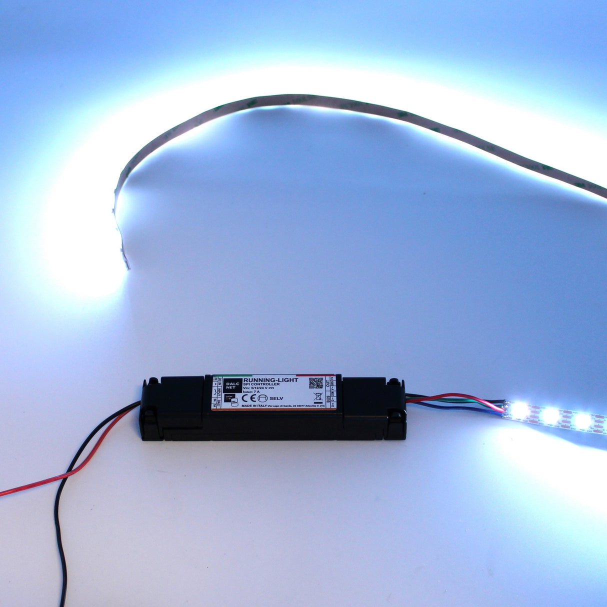 A glowing digital LED strip, powered by a black supply unit with red and black wires on a white surface, emits cool white light—perfect for advanced setups using the SPI Controller for Pixel Digital LED Strip ~ Model RUNNING-LIGHT.