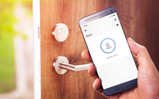 A hand holds a smartphone with the doorBox by Blebox app, remotely unlocking a modern electronic lock on a wooden door. The Smart WiFi Remote Door and Gate Controller shows the lock partially opened.