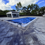 A modern outdoor pool with a blue cover sits on a gray tiled patio featuring Drive-safe Embedding LED Profile ~ Model Dublin XL, enhanced by a glass-walled pool house and trees in the background beneath a partly cloudy sky.