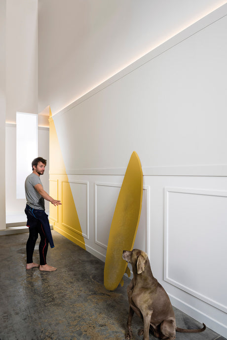 A man gestures toward the Indirect Ceiling Wall Wash LED Profile ~ Model Galicia illuminating a large yellow translucent shape leaning against a white paneled wall, while a brown dog sits on the concrete floor in the modern, brightly lit interior.