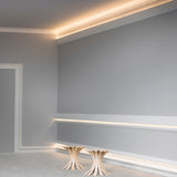 A minimalist room with light gray walls, architectural lighting using the Indirect Ceiling Wall Wash LED Profile ~ Model Galicia along the ceiling, and two small, modern wooden side tables with curved legs.