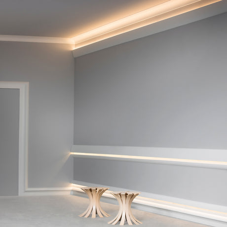 A minimalist room with light gray walls, architectural lighting using the Indirect Ceiling Wall Wash LED Profile ~ Model Galicia along the ceiling, and two small, modern wooden side tables with curved legs.