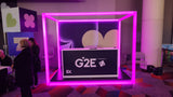 A booth with a G2E sign is framed by a glowing pink neon cube made from 2.91" Square Polycarbonate LED Lighting Tube ~ Model Holston74. People stand nearby on colorful patterned carpet with geometric decorations in the background.