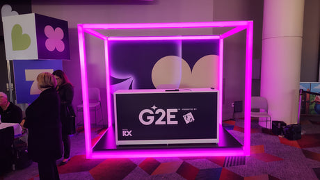 A booth with a G2E sign is framed by a glowing pink neon cube made from 2.91" Square Polycarbonate LED Lighting Tube ~ Model Holston74. People stand nearby on colorful patterned carpet with geometric decorations in the background.