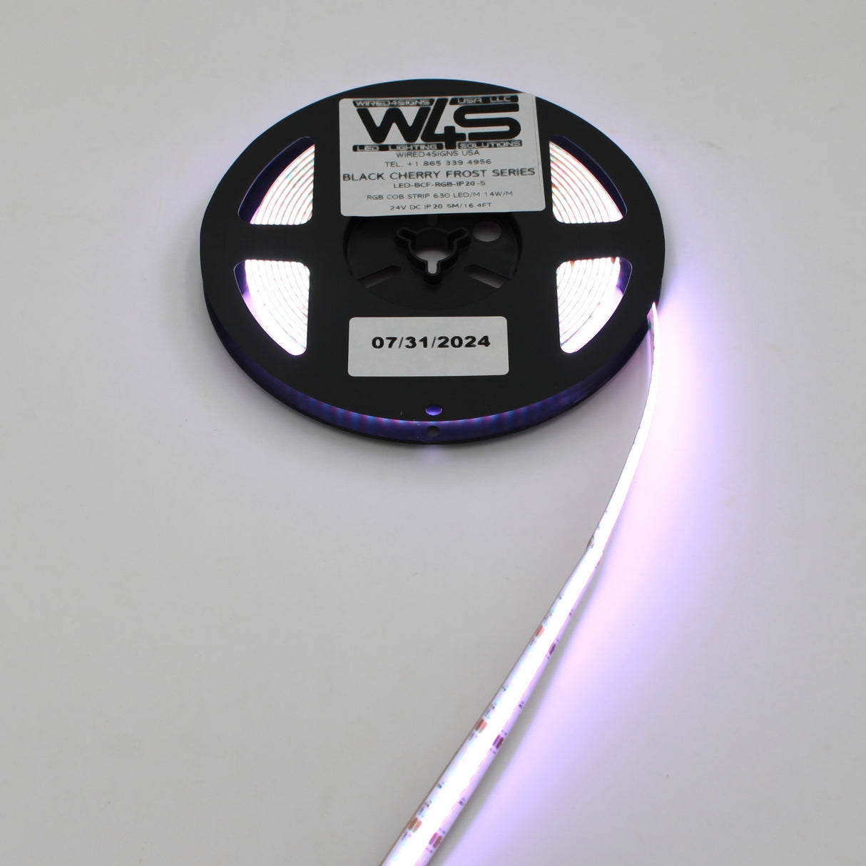 A spool of RGB COB IP20 LED Strip (24V) from the Black Cherry Frost Series glows purple, partially unrolled on a white surface. The reel displays a Black Cherry Frost Series label and a sticker dated 07/31/2024.