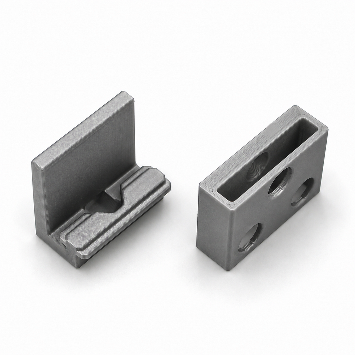 Two machined metal parts: one is the Wall Mounting Bracket for Hannover Profile, an L-shaped bracket with a groove, and the other is a rectangular block with three circular holes and one rectangular opening. Both feature a metallic, polished finish.