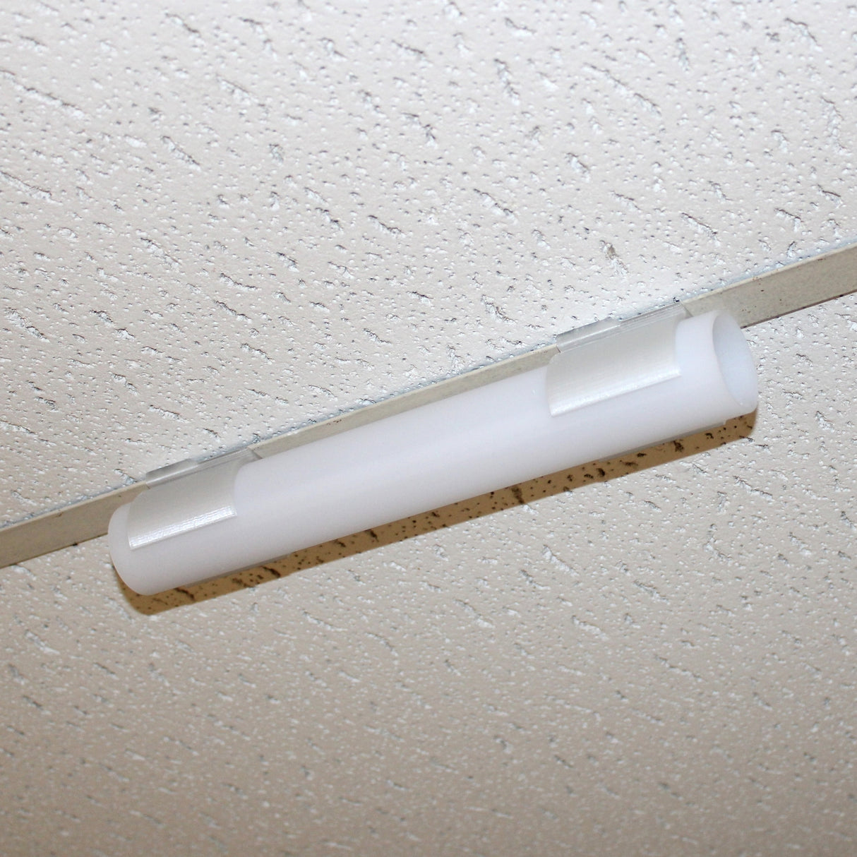 A close-up of a fluorescent light fixture mounted on a white, speckled drop ceiling tile using the Drop Ceiling T-Bar Clip for 1.5 Round Profiles for secure ceiling t-bar installation.
