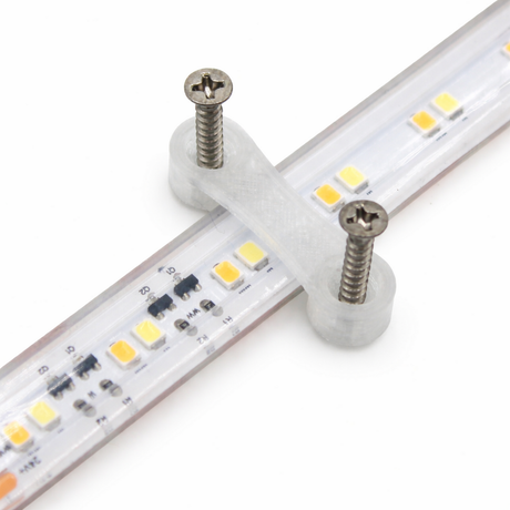 A close-up of the LED Strip Saddle securing a waterproof LED strip to a surface with a clear plastic bracket and two metal screws, showcasing small yellow and white LED chips.