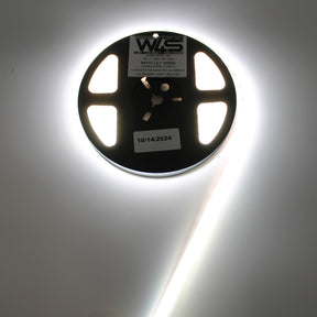 A spool of Continuous COB IP67 LED Strip (24V) ~ White Lily Series glows with smooth, dot-free light. The black reel is labeled 10/14/2024, and the partially unrolled strip shines brightly—ideal for decorative lighting.