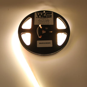 A partially unspooled Continuous COB IP67 LED Strip (24V) ~ White Lily Series glows brightly on a white surface, with a reel labeled “White Lily Series” and a “09/03/2024” sticker—ideal for dot-free decorative lighting.
