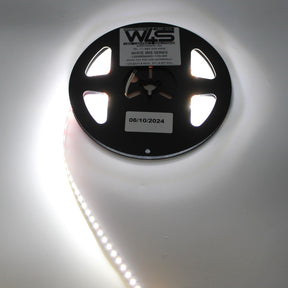 A black reel labeled White Iris Series holds a partially unspooled 24V White High CRI IP20 LED strip with bright white LEDs, lying on a white surface. A sticker on the reel shows the date 06/10/2024.