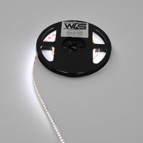 A White High CRI IP20 LED Strip (24V) ~ White Iris Series reel is partially unspooled on a plain white surface, emitting bright light. The labeled reel showcases quality branding, ideal for versatile color temperatures.