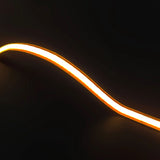 An Ultra-Thin Flexible COB IP20 High CRI LED Strip (24V) ~ EnoLed Micro Series glows with warm orange light, curved into a gentle wave against a dark background.