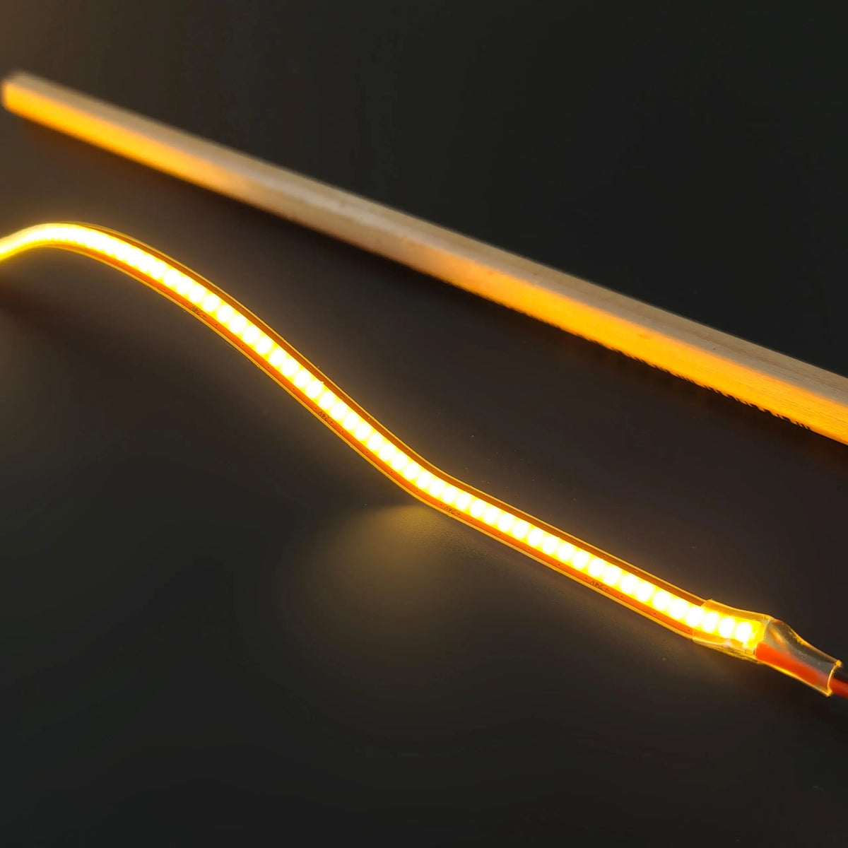 An Ultra-Thin Flexible COB IP20 High CRI LED Strip (24V) ~ EnoLed Micro Series, a flexible, dotless LED strip lying on a dark surface next to a thin wooden rod.