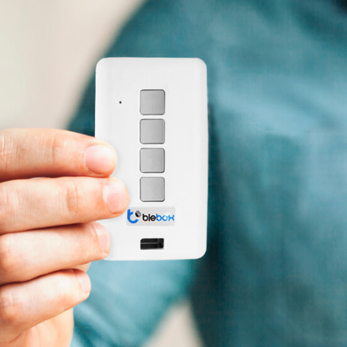 A person holds a Miniature Universal Wi-Fi Remote Control (uRemote by BleBox) with four grey buttons and the blebox logo on the front, featuring micro Wi-Fi technology, against a blurred background.