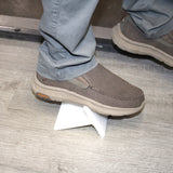 A person wearing gray slip-on shoes and gray pants steps on a small white triangular object—possibly a piece of 2-Part MMA Structural Adhesive for LSE Plastics ~ PE-Bond Kayak500 (50ml 1:1 / B-System)—placed on a wood-patterned floor.