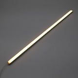 The White Universal Surface-mount Waterproof Outdoor LED Lighting Fixture (24V) ~ Roma Series emits a warm white glow as a slim, dotless LED bar shown diagonally on a plain gray background, with its power cord visible for versatile outdoor use.