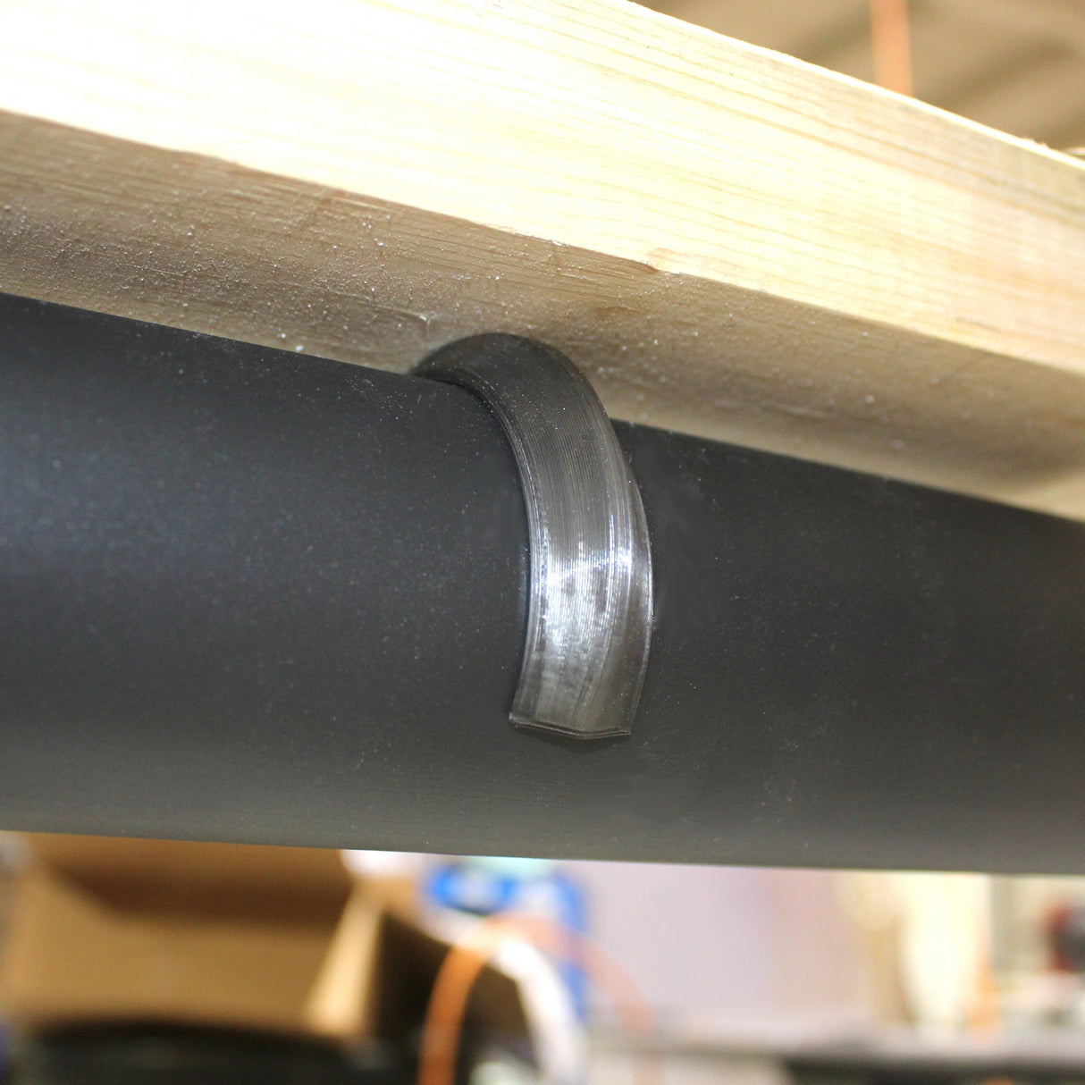 A Mounting Clip for 2.56" Round Profiles secures a black cylindrical pipe to the underside of a wooden surface, next to an LED profile clip, with blurred objects in the background.