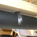 A Mounting Clip for 2.56" Round Profiles secures a black cylindrical pipe to the underside of a wooden surface, next to an LED profile clip, with blurred objects in the background.
