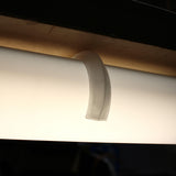 A Mounting Clip for 2.56" Round Profiles secures a glowing fluorescent light under a wooden shelf with its curved, translucent plastic design.