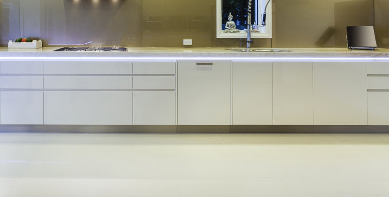 Modern kitchen with glossy white cabinets, Smart10 Miniature Surface Mount LED channel under-cabinet lighting, gas cooktop on the left, sink beneath a window in the center, beige tile flooring, and vegetables with a small appliance on the countertop.