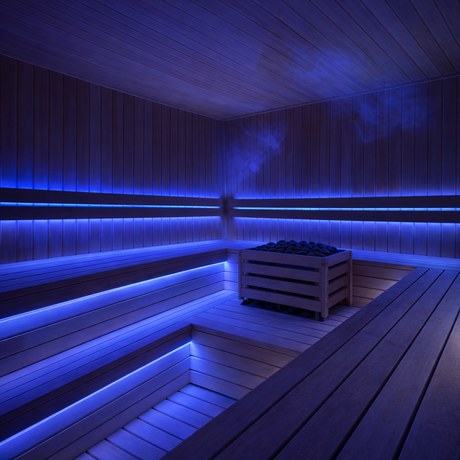 A modern sauna room with wooden benches and walls, lit by a Sauna Pro RGB Series—RGB IP68 Waterproof LED Strip (24V)—casting blue light, with faint steam rising from heated stones in a central heater.