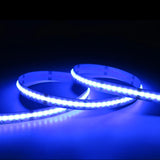 The RGB+CCT COB IP20 High CRI LED Strip (24V) ~ EnoLED UniColor Series emits a vivid blue glow, coiled in loops against a dark background and brightly illuminating the surrounding area.