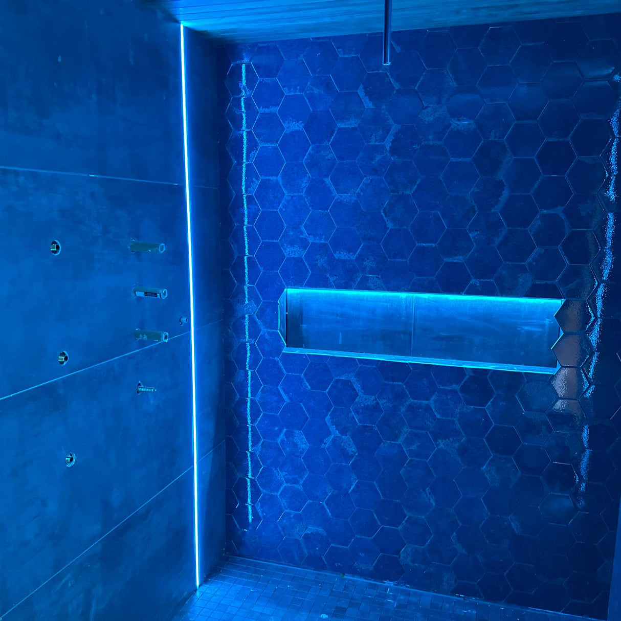 A modern shower featuring blue hexagonal tiles and a built-in shelf illuminated by the Tile Edge LED Strip Light Channel ~ Model Uni-Tile12 180Deg, complemented by sleek metal fixtures on the left wall to create a clean, futuristic atmosphere.