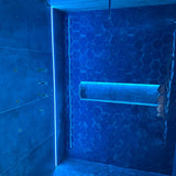 A modern shower featuring blue hexagonal tiles and a built-in shelf illuminated by the Tile Edge LED Strip Light Channel ~ Model Uni-Tile12 180Deg, complemented by sleek metal fixtures on the left wall to create a clean, futuristic atmosphere.