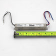 A Slimline Small Constant Voltage LED Driver (24V) from the ERP Power VLM Series, with wires at both ends, is placed above a yellow measuring tape showing its length of about 6 inches (15 cm); specs and constant voltage are shown on its label.