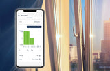 A smartphone shows the openSensor by Blebox app’s history bar chart for an open window, with a bright cityscape and sunlight streaming into the room.