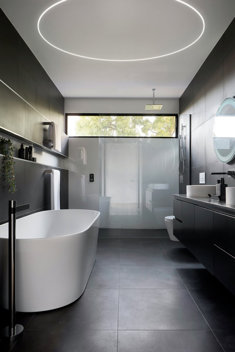 Modern bathroom with dark tiles, freestanding tub, floating black vanity, round backlit mirror, open glass shower, large window, and 0.4 Bendable Drywall LED Lighting Channel (Klus KOZEL-10-BD) for curved linear lighting and minimalist style.