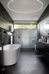 Modern bathroom with dark tiles, freestanding tub, floating black vanity, round backlit mirror, open glass shower, large window, and 0.4 Bendable Drywall LED Lighting Channel (Klus KOZEL-10-BD) for curved linear lighting and minimalist style.