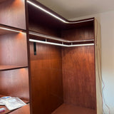 A wooden closet features the Lighted Closet Rod LED Channel ~ Model Alu-Round with an aluminum LED profile on shelf and top edges, a hanging rod supported by a black bracket, and open shelving on the left showing visible wires and packets.