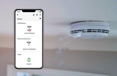 A smartphone displays the inputSensorQ by Blebox alerting to smoke detected, while a ceiling-mounted smoke detector emits visible smoke—demonstrating how the 4-Channel Smart Wi-Fi Input Status Module enhances home safety automation.