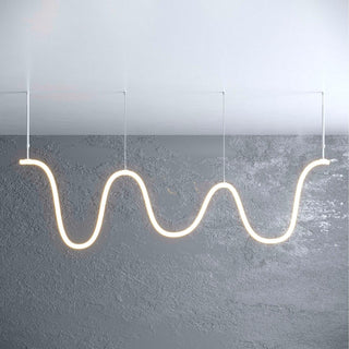 An Adjustable Suspension Arm for the Neon 360 Series supports a wavy, glowing tube pendant light, hanging elegantly from the ceiling against a textured gray wall and ceiling backdrop.