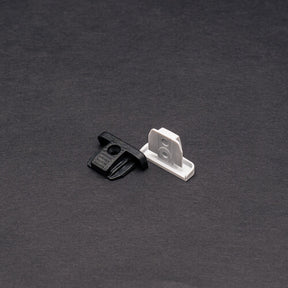 Two small plastic mounting clips, one black and one white, are shown on a dark surface. Designed for the End Cap for Power Track Twist, they have flat bases and upright tabs with center holes for secure attachment.
