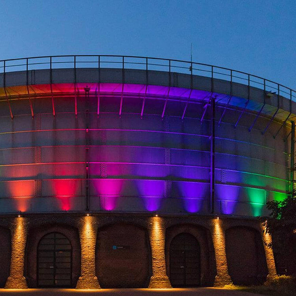 A large round brick building with arched doorways is illuminated at dusk by Color-Changing Outdoor LED Wall Washer Lights (Revo ColorFlow Casambi Series), casting vibrant rainbow hues against the evening sky.