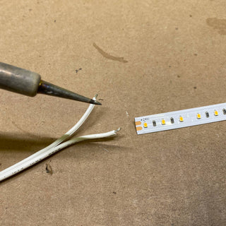 A close-up of a Solder Joint - 2 conductors, with a soldering iron aimed at two stripped white wires and a cut LED strip on a brown work surface, ready for precise soldering.