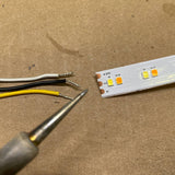 Close-up of expert soldering on Solder Joint - 3 conductors (white, black, yellow wires) and an LED strip labeled “+24V” and “W” on a brown surface—ideal for quality solder joint work.