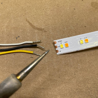 Close-up of expert soldering on Solder Joint - 3 conductors (white, black, yellow wires) and an LED strip labeled “+24V” and “W” on a brown surface—ideal for quality solder joint work.