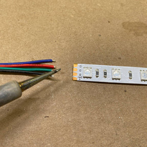The Solder Joint - 4 conductors is shown with a soldering iron near four colored wires (black, blue, green, and red) and LED strip connector pads on a brown surface, ready for precise soldering.