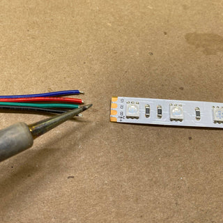 The Solder Joint - 4 conductors is shown with a soldering iron near four colored wires (black, blue, green, and red) and LED strip connector pads on a brown surface, ready for precise soldering.