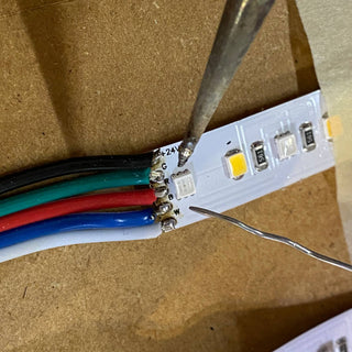 Close-up of a Solder Joint - 5 conductors being used to securely connect black, green, red, blue, and white wires to an LED strip’s circuit board on a wooden surface.