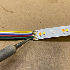 A close-up of a Solder Joint - 6 conductors, showing a soldering iron forming a clean connection on multicolored wires attached to an LED strip on a brown surface.