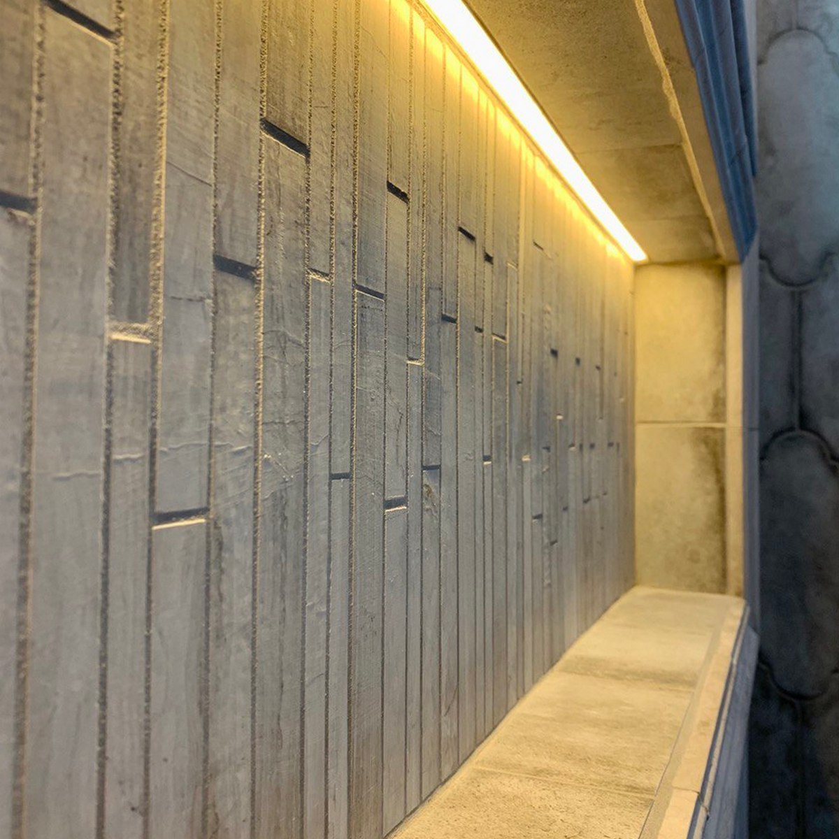 Close-up of a textured concrete wall with vertical lines, illuminated along the top by the Tile Edge LED Strip Light Channel ~ Model Uni-Tile12 180Deg. The warm yellow LED accent lighting from this channel enhances the sleek, modern appearance and adds depth to the minimalist architectural design.